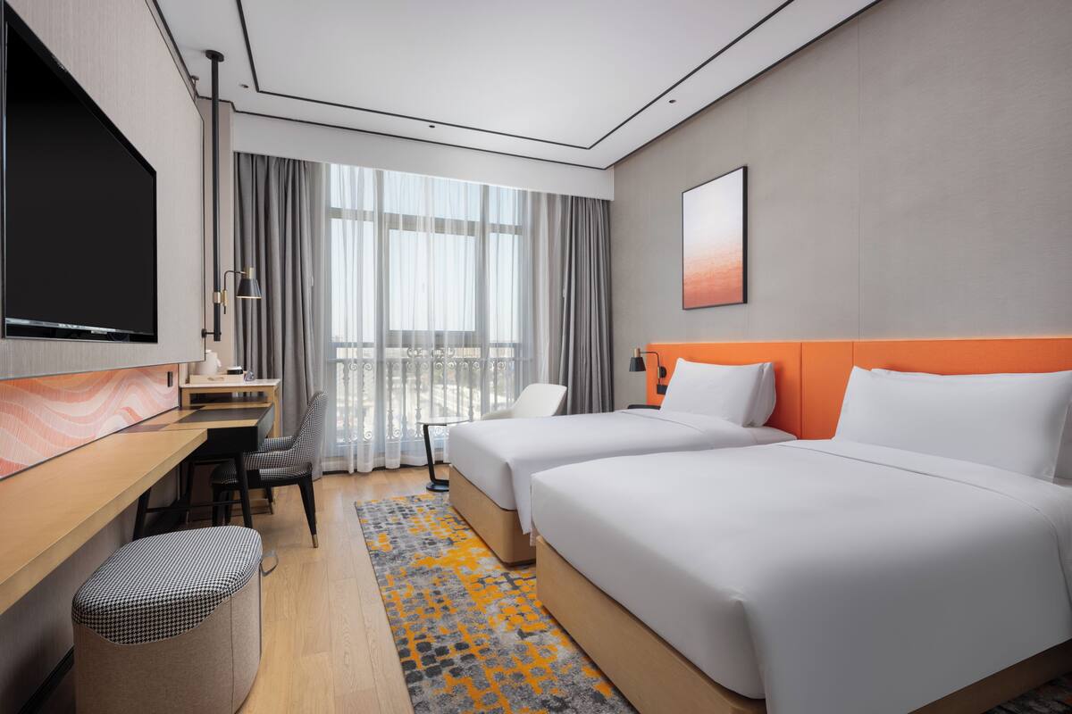 Habitación de hotel con dos camas, un escritorio, televisor y un ventanal con cortinas.
