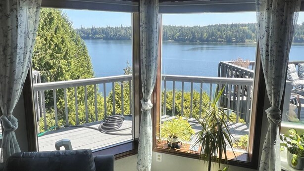 A room with a large window overlooking a lake and forest. There is a balcony with a railing and potted plants. - Waterfront charming 3-bedroom house with hot tub, sauna and bikes, sleeps 6 (Shelton)