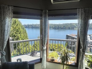 Et værelse med stort vindue der kigger ud over en sø og skov. Der er en balkon med gelænder og plantekasser.