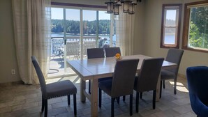 A dining area with a table and chairs, large windows with a view of a lake and forest, and a chandelier. - Waterfront charming 3-bedroom house with hot tub, sauna and bikes, sleeps 6 (Shelton)