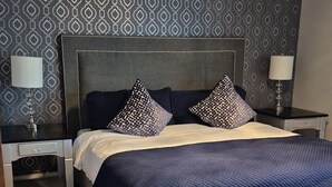 A modern bedroom with a large bed, two nightstands, a bench, patterned wallpaper, and a wooden floor. - Waterfront charming 3-bedroom house with hot tub, sauna and bikes, sleeps 6 (Shelton)