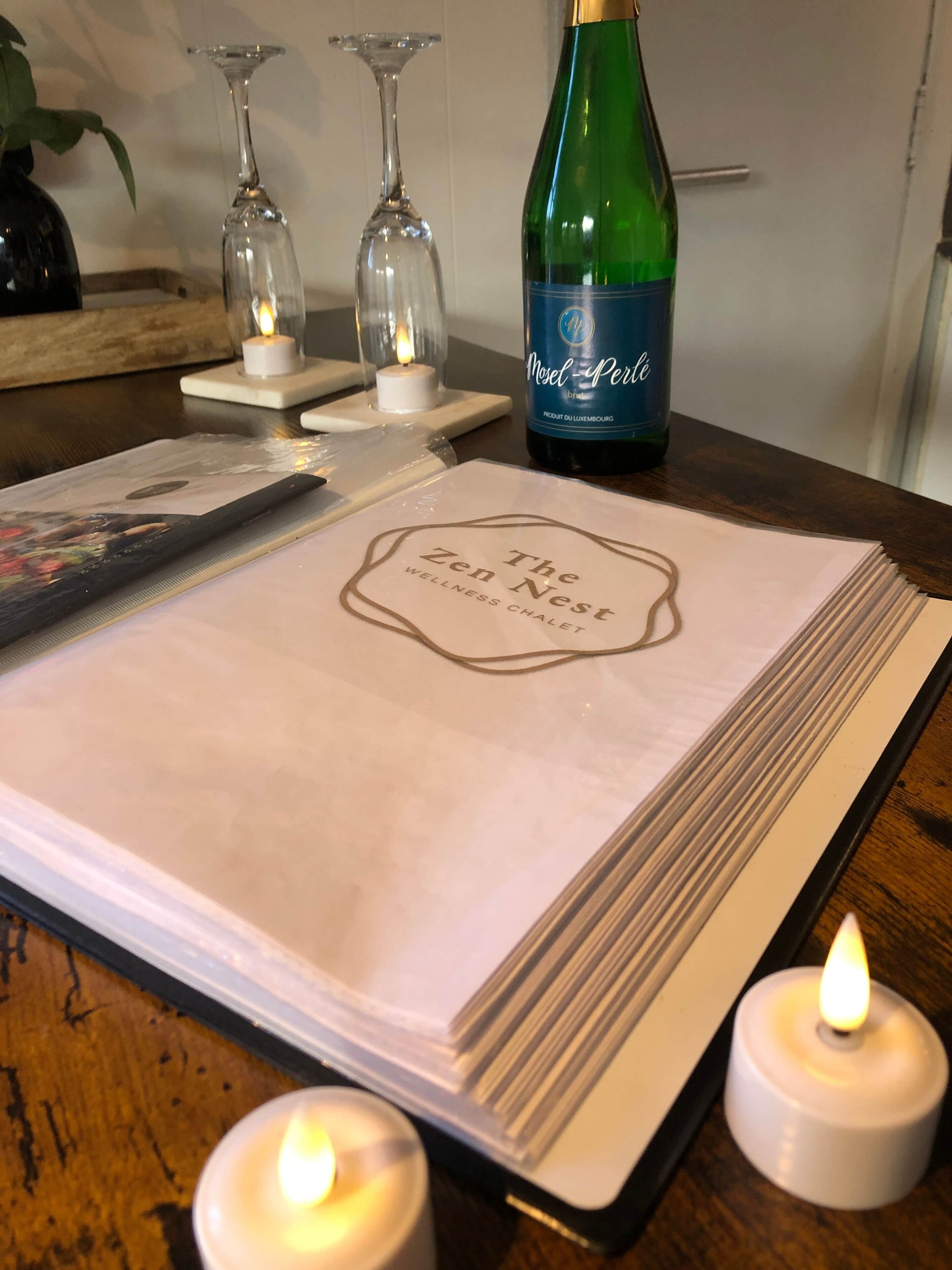 A table with a placemat labeled "The Zen Nest Wellness Chalet," a bottle of Miel Perle, two glasses, and two lit candles.