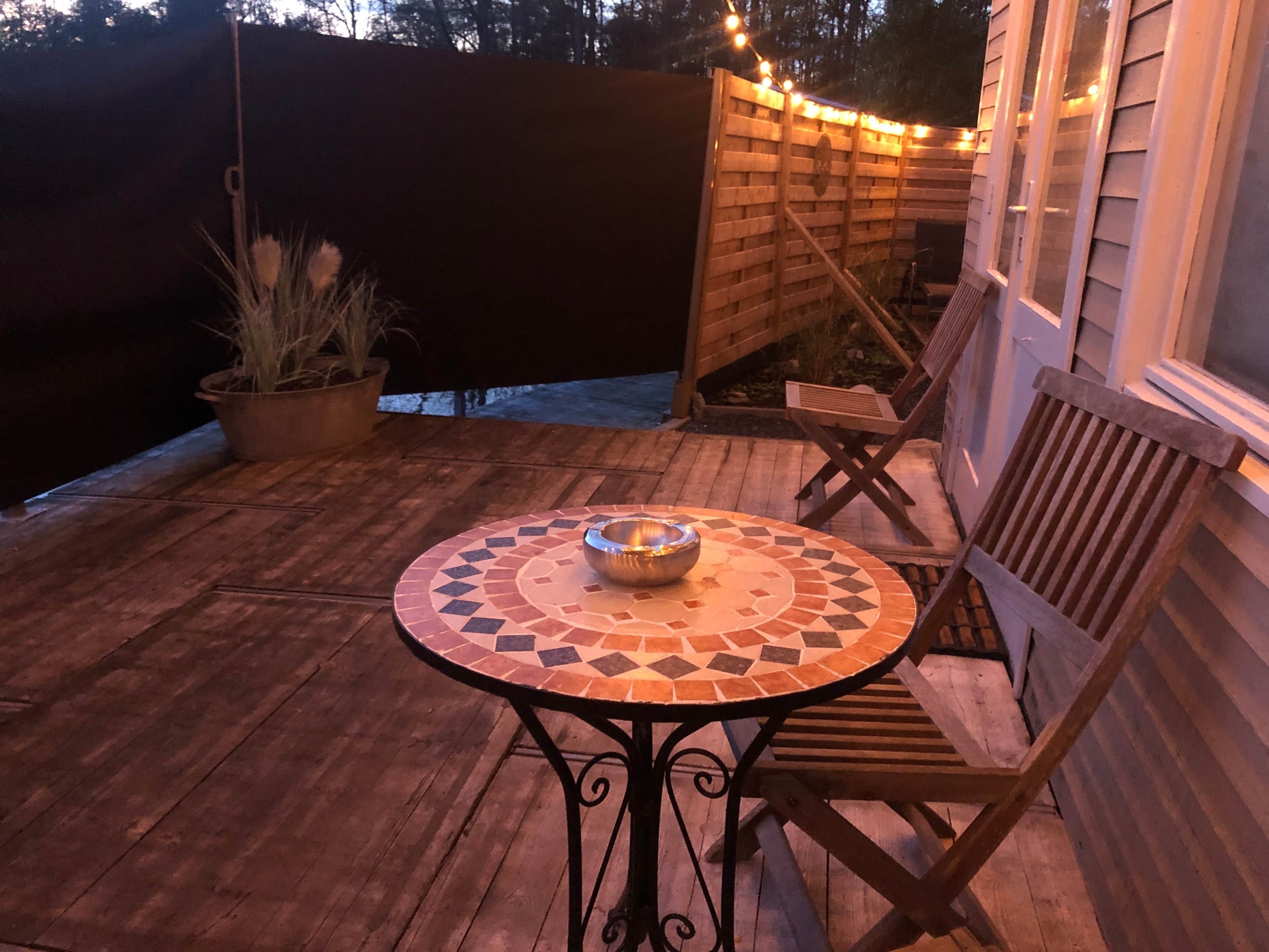 Een houten terras met een mozaiektafel en stoelen, een potplant en lichtjes.