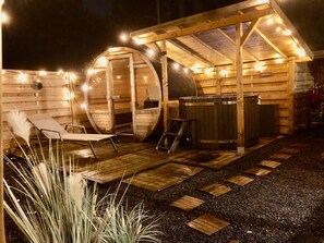 A wooden spa structure with a hot tub and a lounge chair, surrounded by string lights.