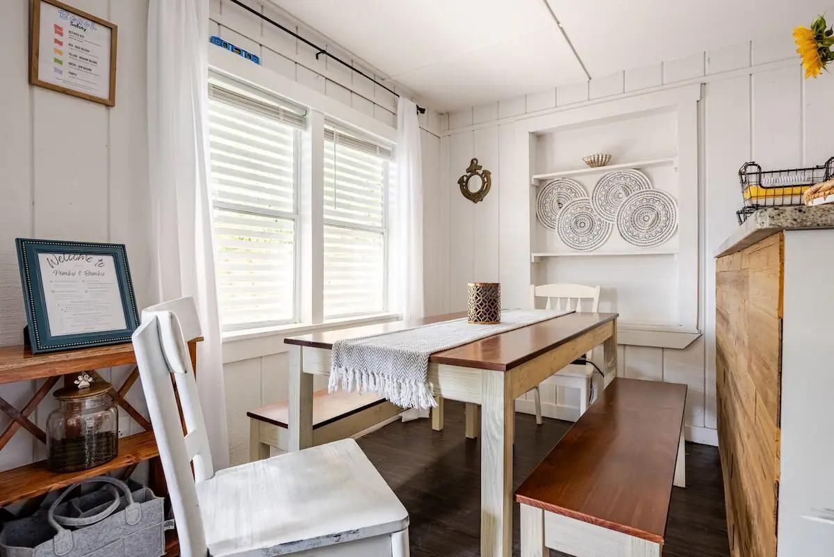 A dining area with a wooden table and chairs, a white wall with shelves holding plates and a lantern, and a window with blinds.