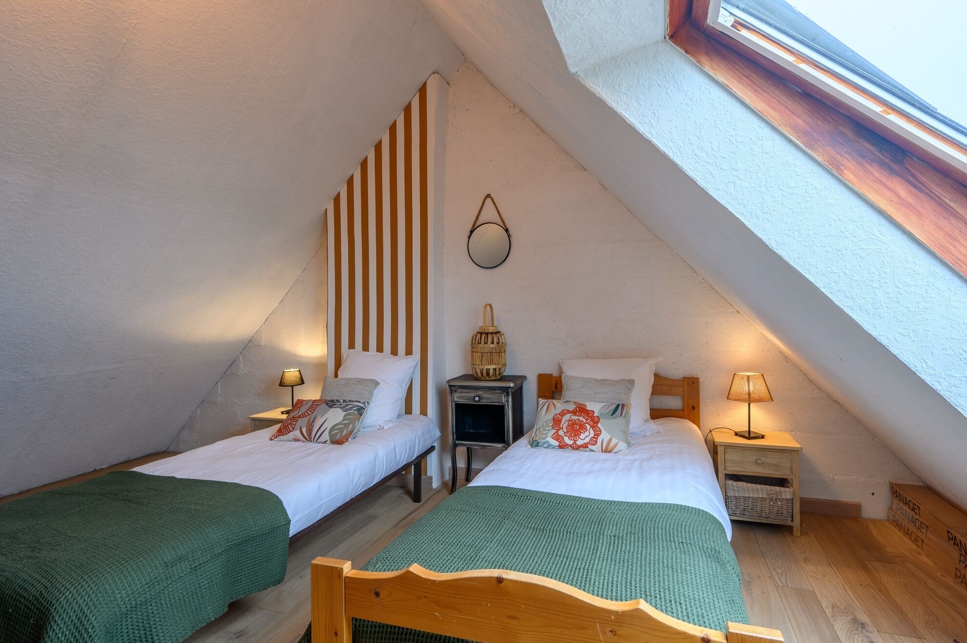 Une chambre mansardée accueillante, avec deux lits simples, une petite table, une lampe et une fenêtre avec un cadre en bois.