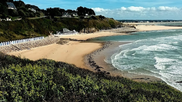 Una playa costera con costa rocosa, una playa de arena y una hilera de cabañas de playa blancas.