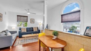 Una sala de estar con un sofá, una mesa de centro y una mesa de comedor con sillas. Se ve una ventana con persianas y un ventilador de techo.