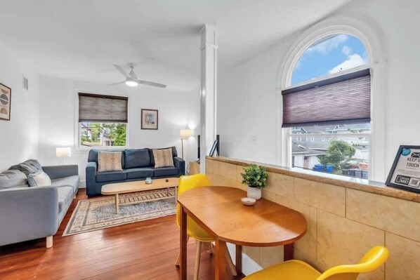 A living room with a sofa, a coffee table, and a dining table with chairs. There is a window with blinds and a ceiling fan.