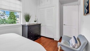 Un dormitorio con una cama, un tocador, un sillón gris y armarios blancos.