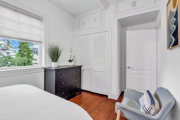 Un dormitorio con una cama, un tocador, un sillón gris y armarios blancos.
