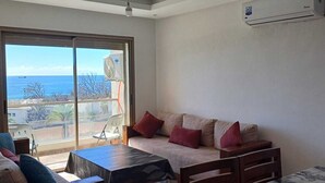 A room with a sofa, a table, and a chair. There is a large window with a view of the sea and buildings. An air conditioning unit is mounted on the wall. - Sea House Agadir (Agadir)