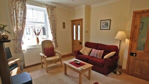 A living room with a red sofa, a wooden coffee table, a white armchair, and a red armchair. There is a window with curtains, a lamp, and a framed picture on the wall. - 2 bedroom accommodation in Warkworth (Warkworth)