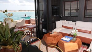 A modern interior with a sofa, coffee table, and potted orchid. A boat is visible outside the window. - House of Hütter - Floating Home Goldfinger (Formentera)