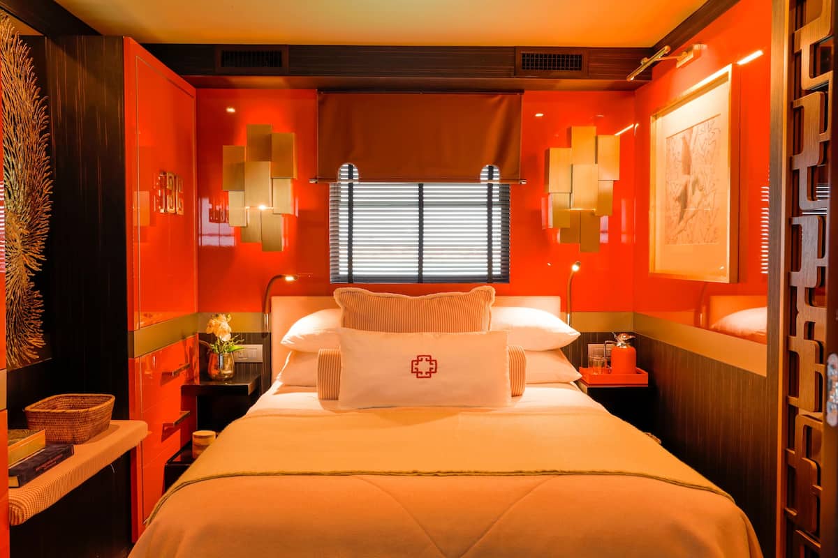 Une chambre d’hôtel avec un grand lit, une table de chevet avec une lampe, une fenêtre avec des stores et une œuvre d’art accrochée au mur.