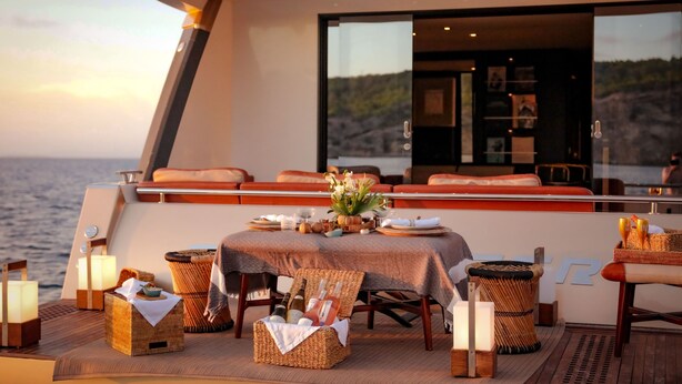 A dining area on a boat with a table set for two, surrounded by wicker baskets and candles. - House of Hütter - Floating Home Goldfinger (Formentera)