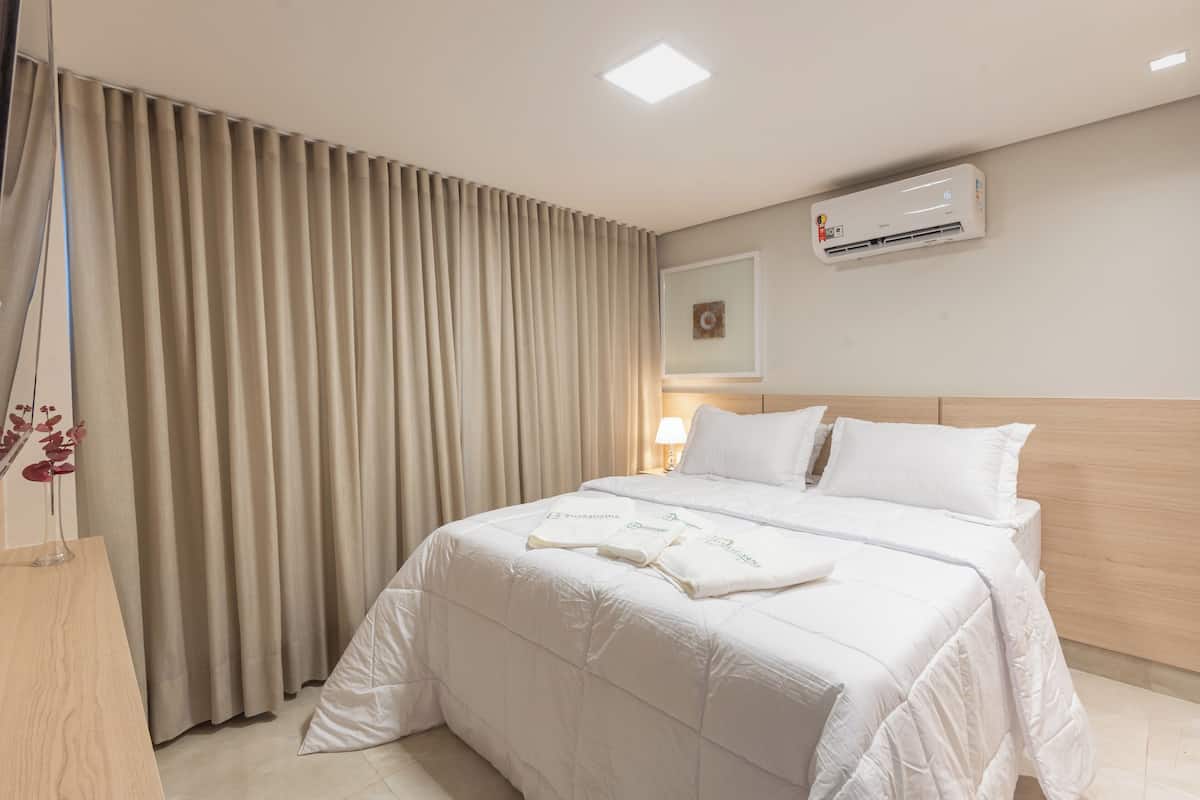 A hotel room with a bed, beige curtains, an air conditioning unit, and a bedside lamp.