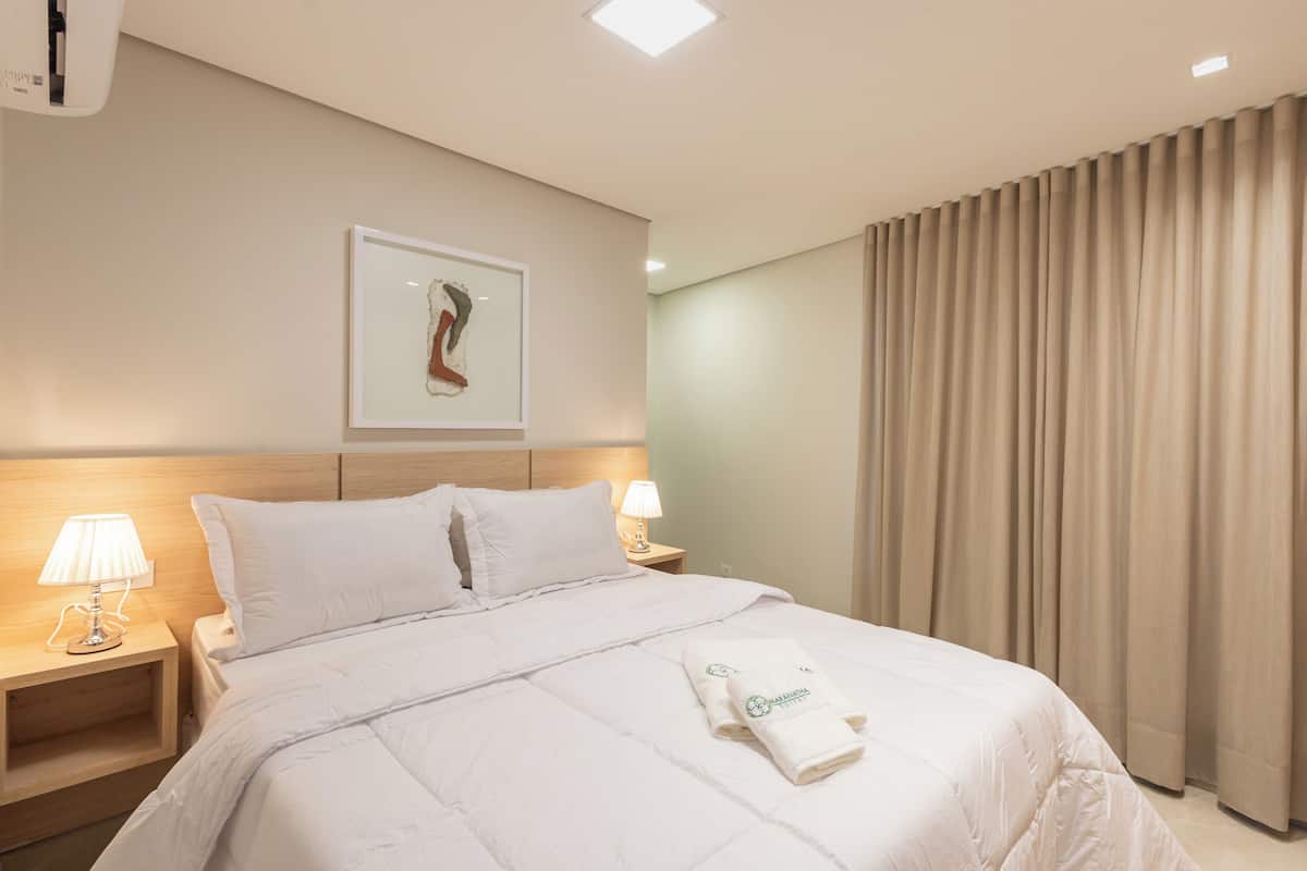 A hotel room with a neatly made bed, a framed artwork on the wall, and a window with curtains.