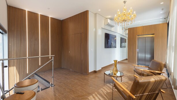 A modern hotel lobby with a wooden floor, a chandelier, a seating area with leather chairs, and a staircase with a glass railing. - Maranatha Suítes (Campo Grande)