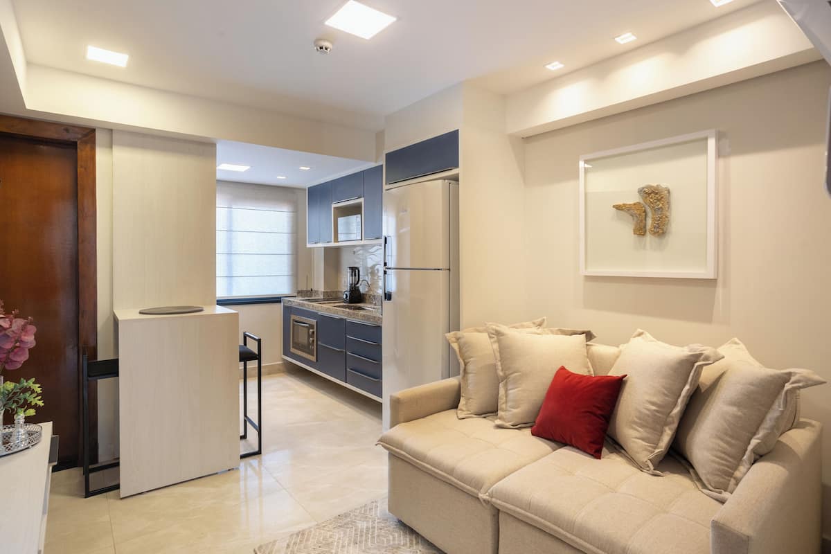 A modern living room with a beige sofa, a small coffee table, and a red pillow. There is a kitchen area with a refrigerator, a sink, and cabinets.