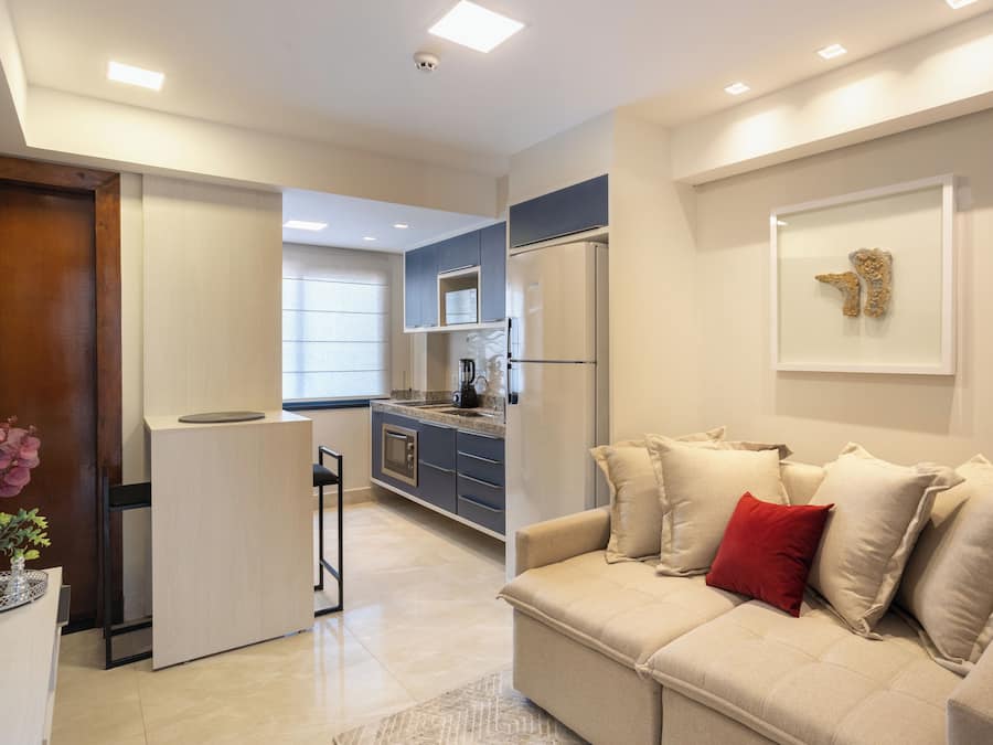 A modern living room with a beige sofa, a small coffee table, and a red pillow. There is a kitchen area with a refrigerator, a sink, and cabinets.