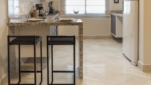 A modern kitchen with a marble countertop, two bar stools, a coffee maker, and a refrigerator. - Maranatha Suítes (Campo Grande)