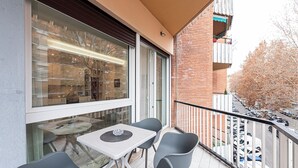Uma varanda com mesa e cadeiras, uma porta de vidro e vista para carros estacionados e árvores.