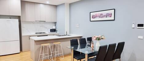 Une cuisine moderne avec des meubles blancs, un four à micro-ondes et une cuisinière à gaz. On y trouve également un coin repas avec une table en verre et des chaises noires.