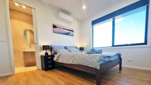 Ein Schlafzimmer mit Holzboden, einem Bett mit gemustertem Bettbezug, einem Nachttisch mit Lampe, einer Wand-Klimaanlage, einem Fenster mit Jalousien und einem Bild an der Wand.