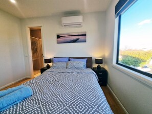 A bedroom with a bed, two bedside tables with lamps, an air conditioning unit, a painting, and a window with a view. - Rosie Townhouse - Modern and Comfortable Home (Sunshine North)