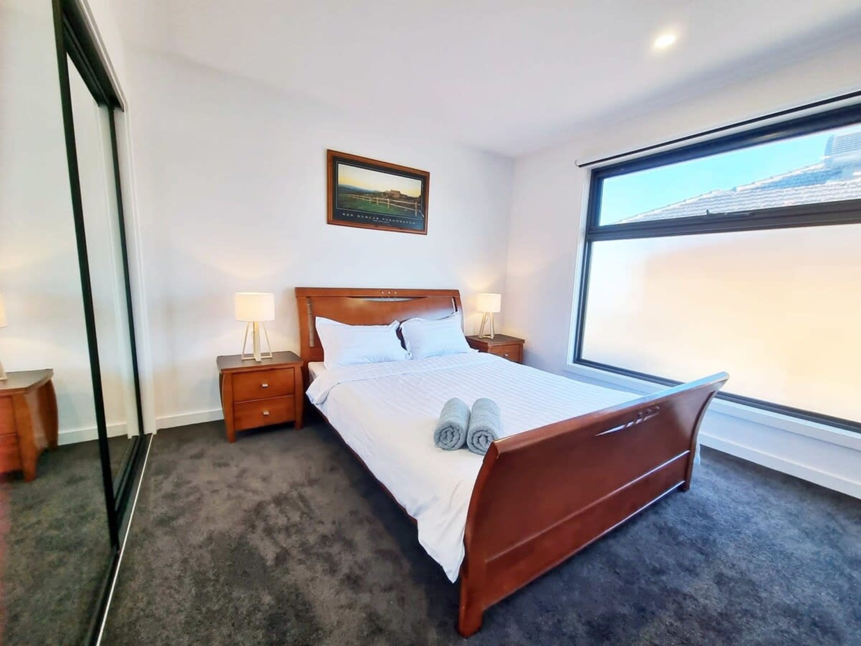 A bedroom with a wooden bed, two bedside tables with lamps, a framed picture on the wall, and a large window.