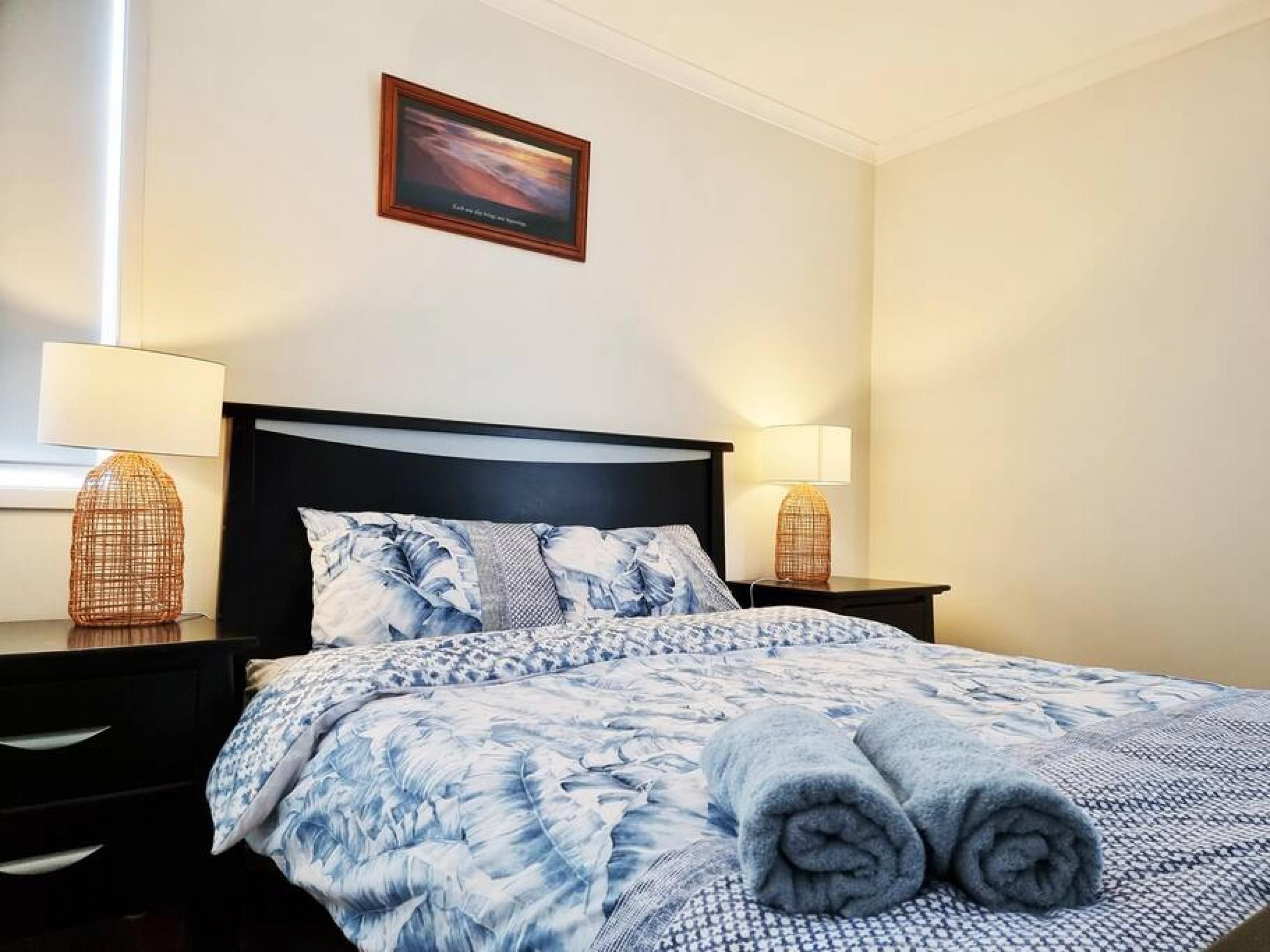 Una cama bien hecha con una colcha azul y blanca estampada, dos lámparas de noche y un cuadro en la pared.