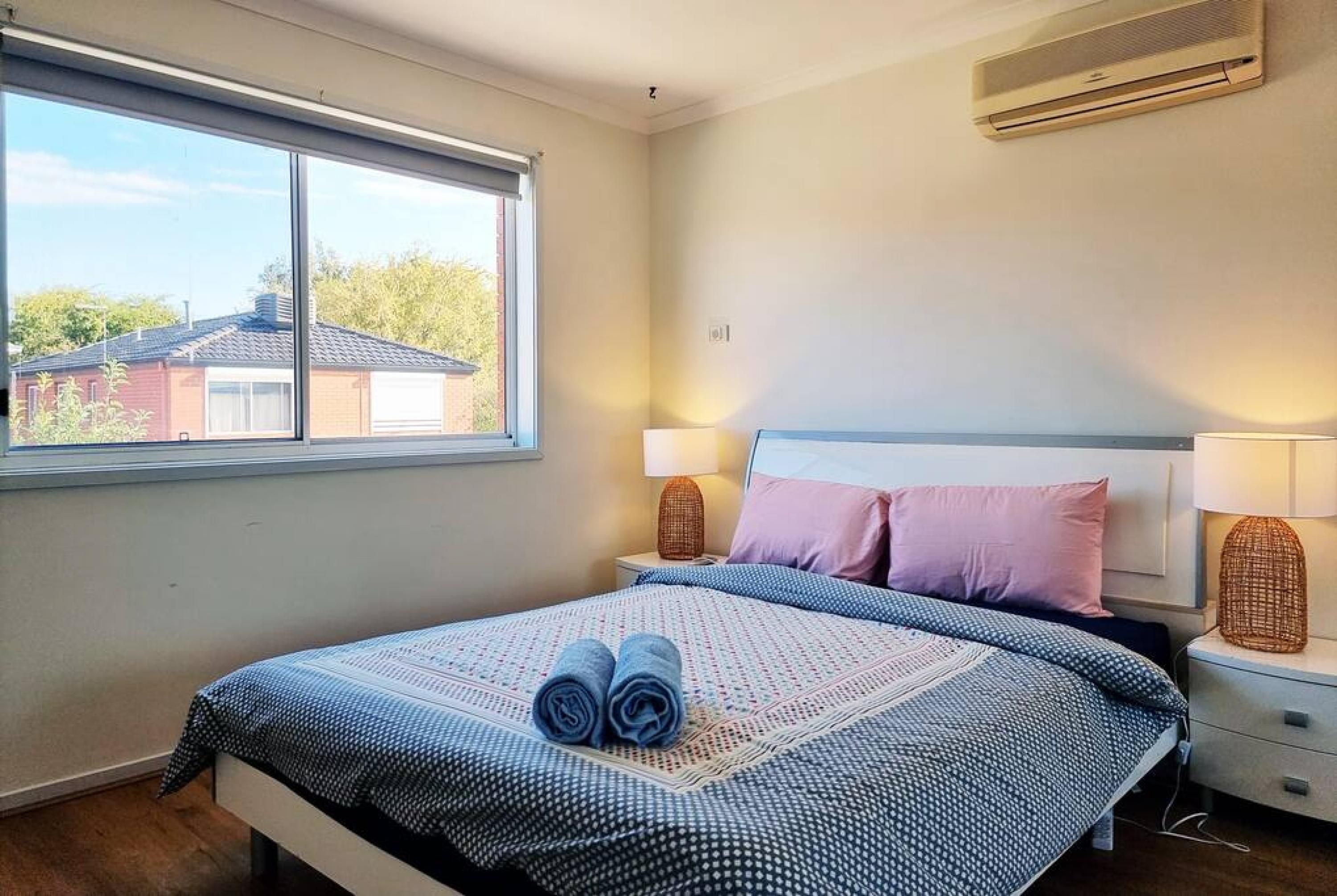 A bedroom with a bed, two bedside lamps, a window, and an air conditioning unit.