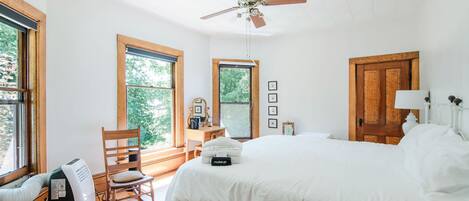 A bedroom with a large bed, a ceiling fan, wooden flooring, and windows with views of greenery.