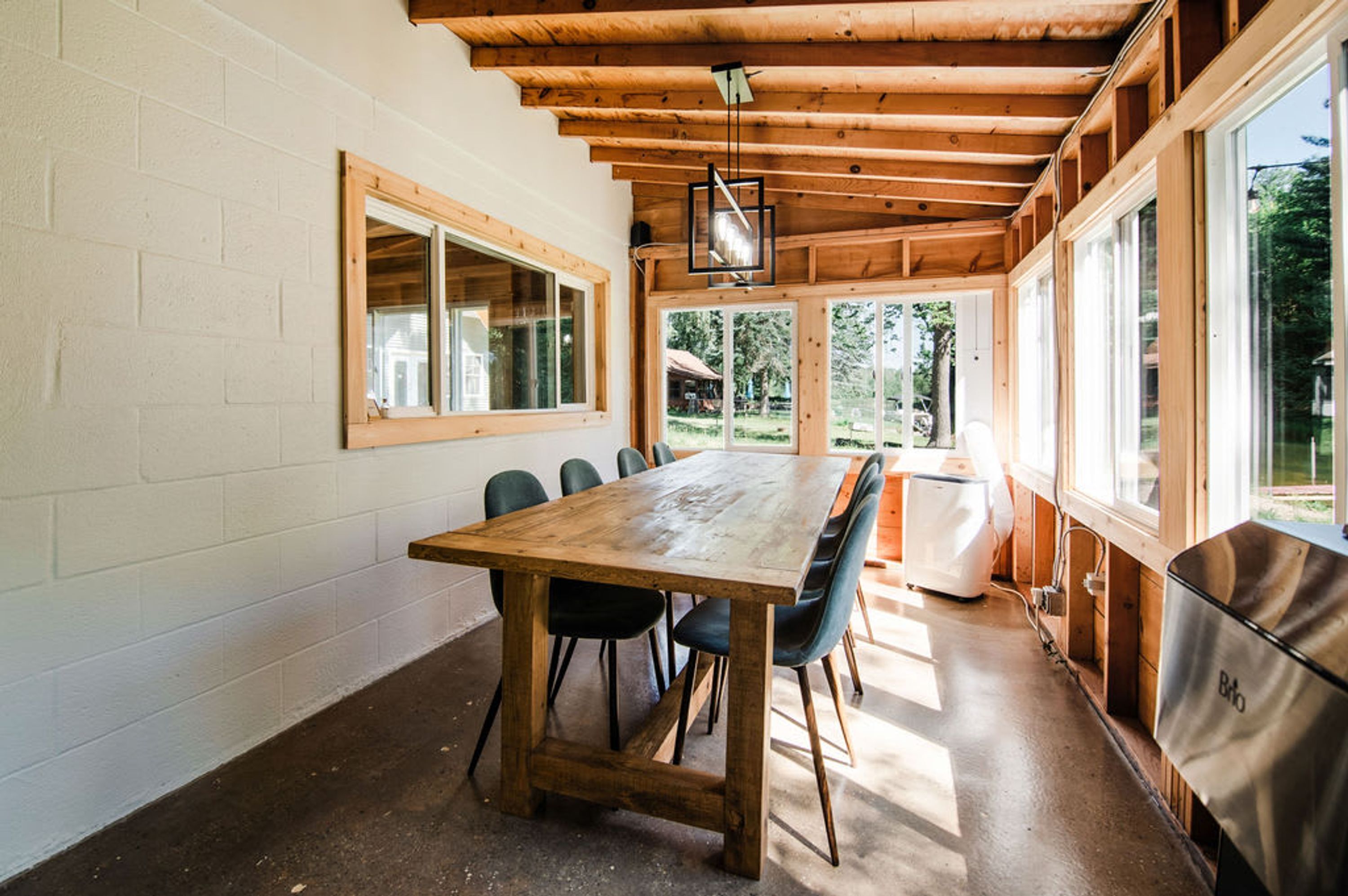 Une table à manger en bois, des chaises, un luminaire suspendu et de grandes fenêtres.