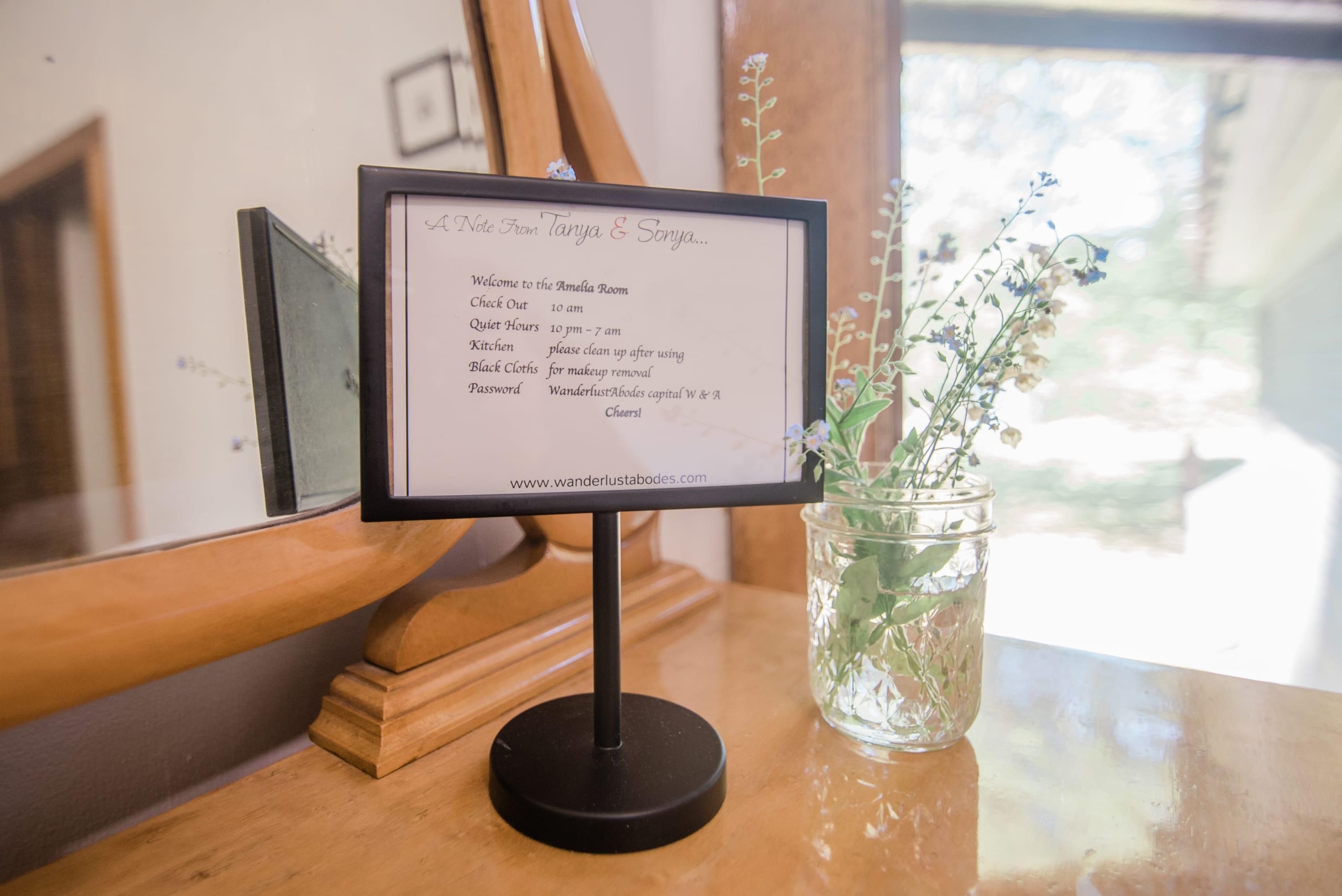Un letrero con información de las habitaciones sobre un atril, una mesa de madera y un jarrón de vidrio con flores.