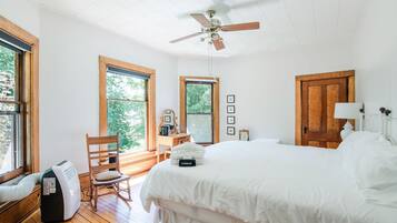 A bedroom with a large bed, a ceiling fan, wooden flooring, and large windows.