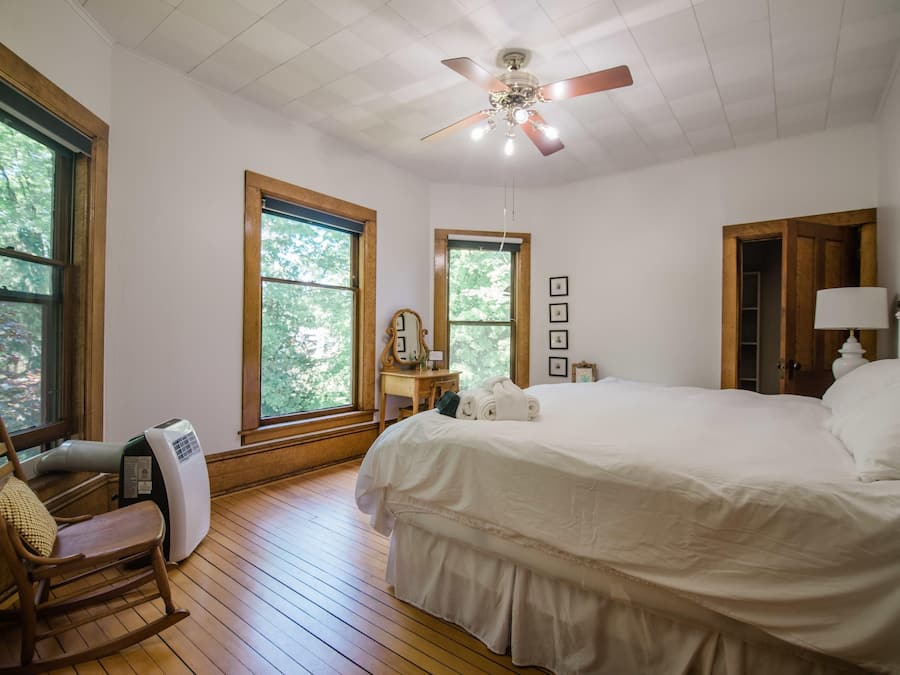 Un dormitorio con una cama grande, suelo de madera, ventilador de techo y ventanas amplias.
