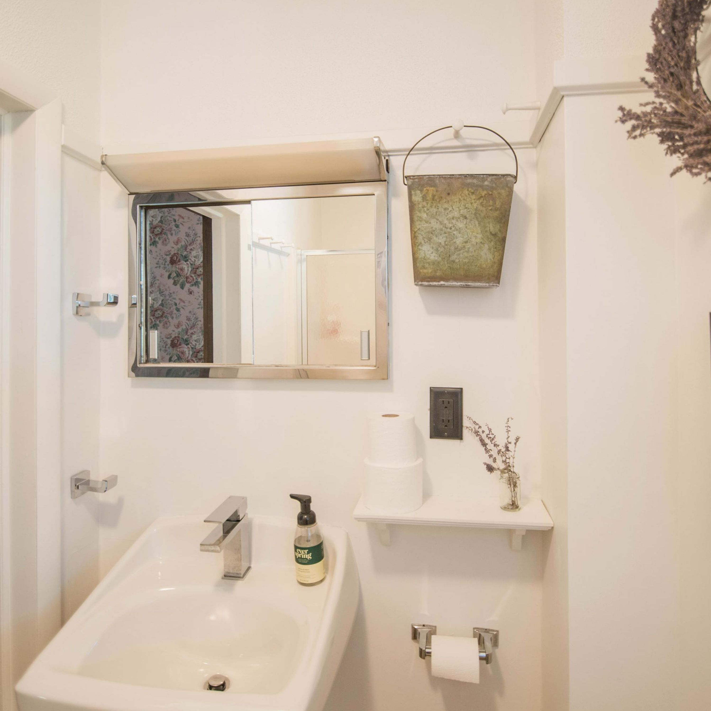 Un baño con lavamanos, espejo, dispensador de papel higiénico y una pequeña estantería con un florero.