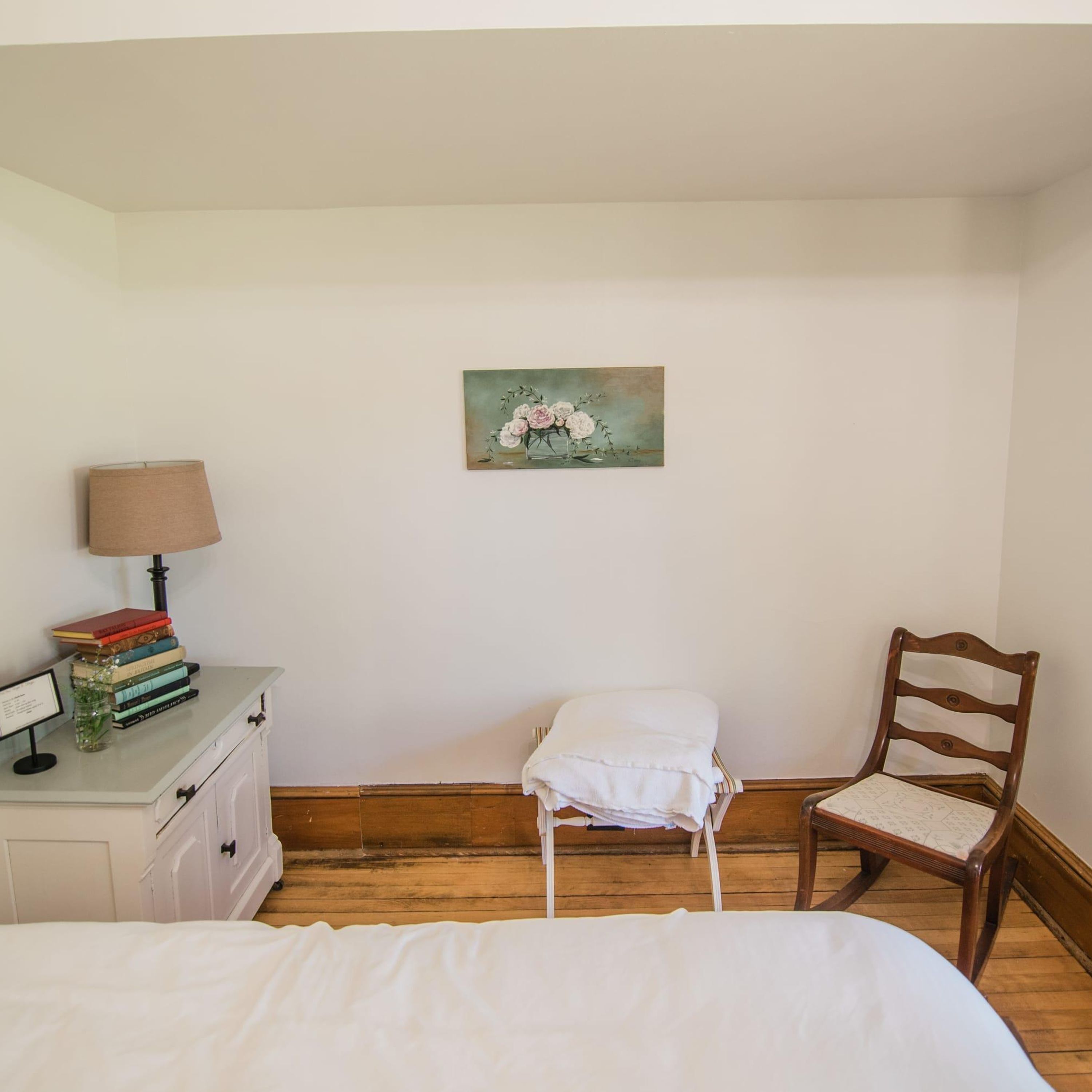 Un dormitorio con cama, mesita de noche con lámpara, una silla y un cuadro en la pared.