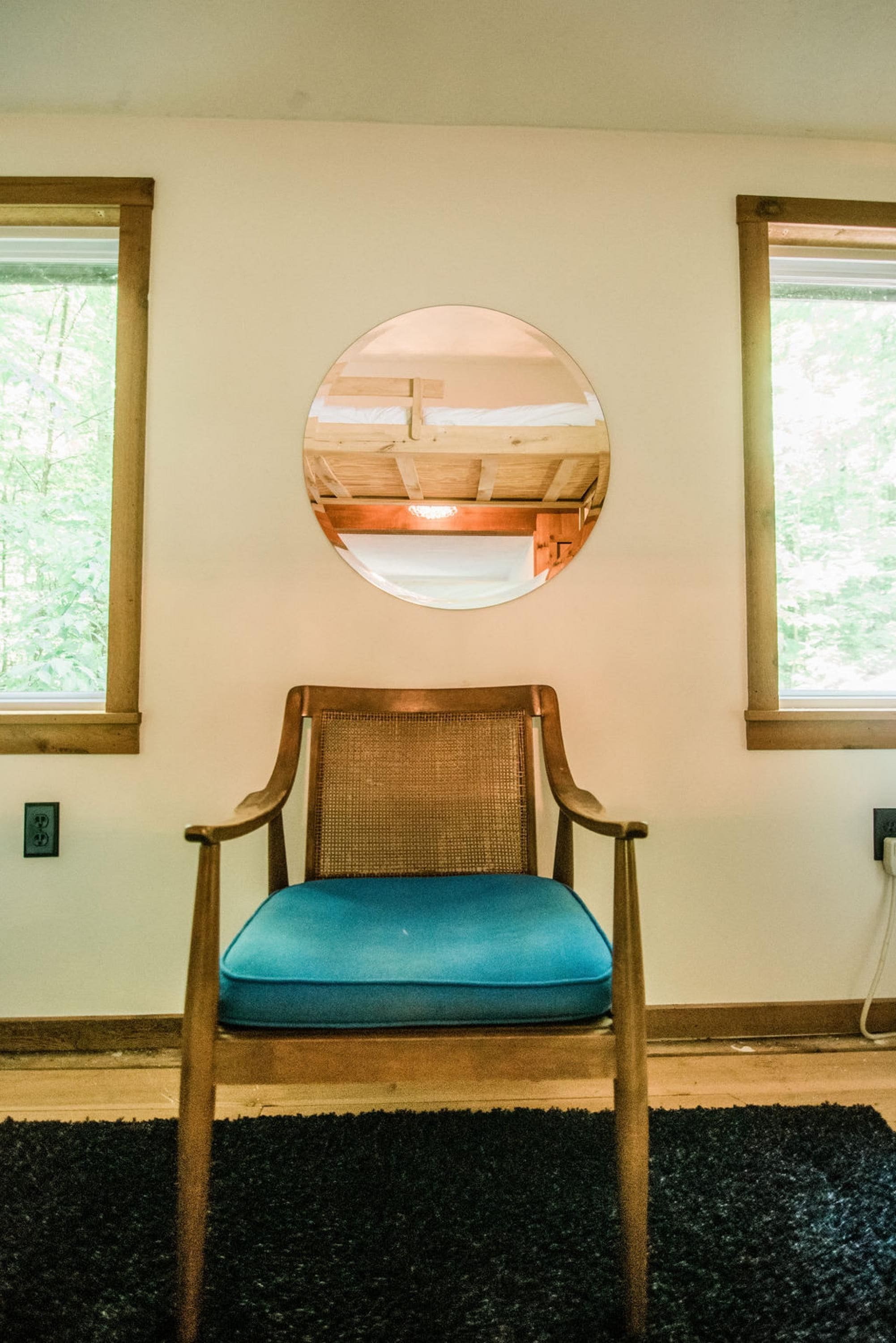 A wooden chair with a blue cushion in a room with two windows and a round mirror on the wall.