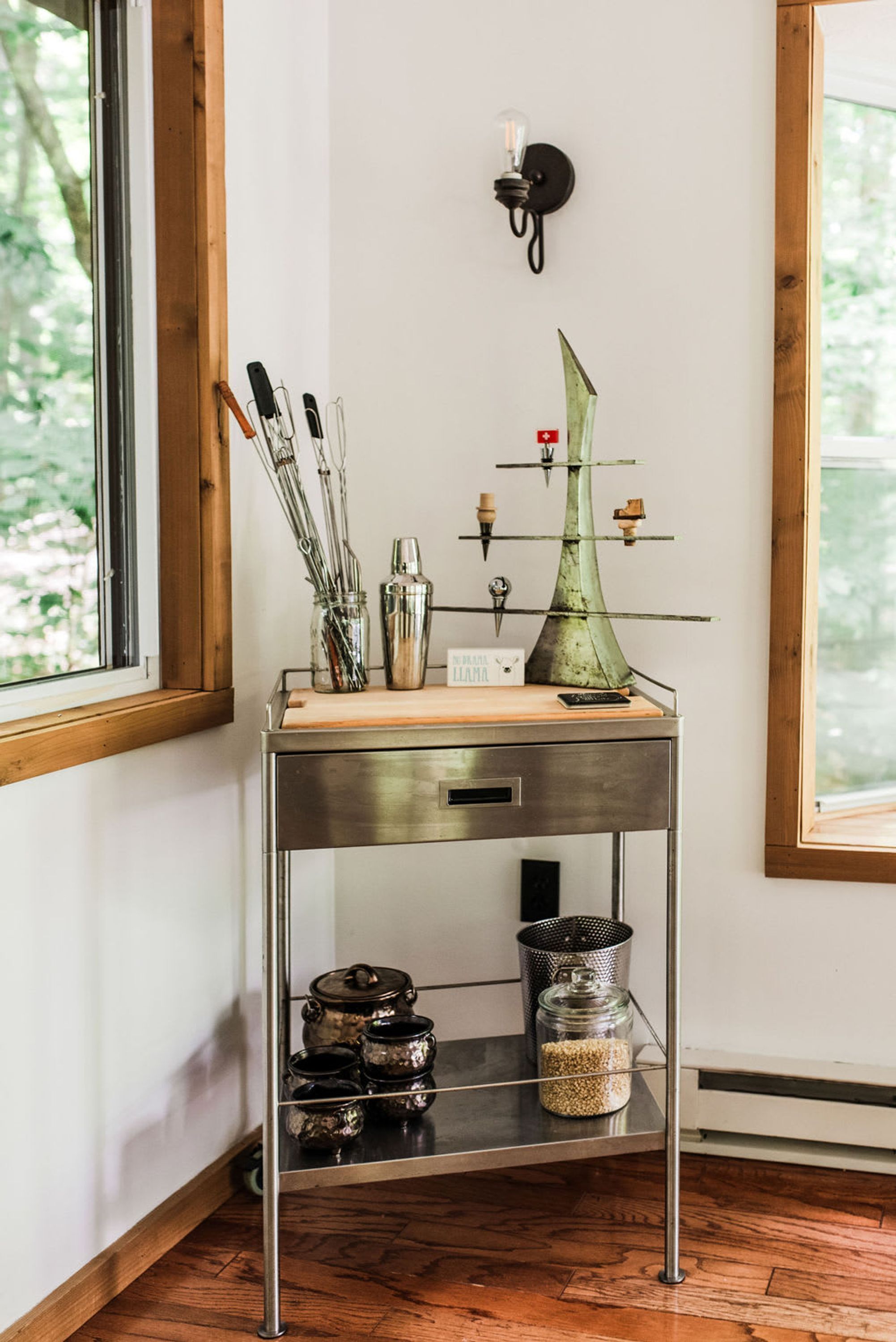 Esquina de una habitación con un marco de ventana de madera, una mesa metálica con utensilios de cocina, un adorno verde y una lámpara fijada a la pared.
