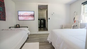 Ein Schlafzimmer mit zwei Betten, einem Fernseher an der Wand und einer durch eine offene Tür einsehbarer Küche.
