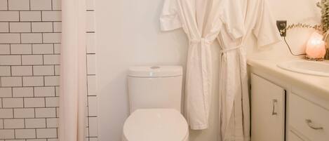 Une salle de bain avec des toilettes blanches, une douche avec un rideau blanc et deux peignoirs blancs suspendus à des crochets.