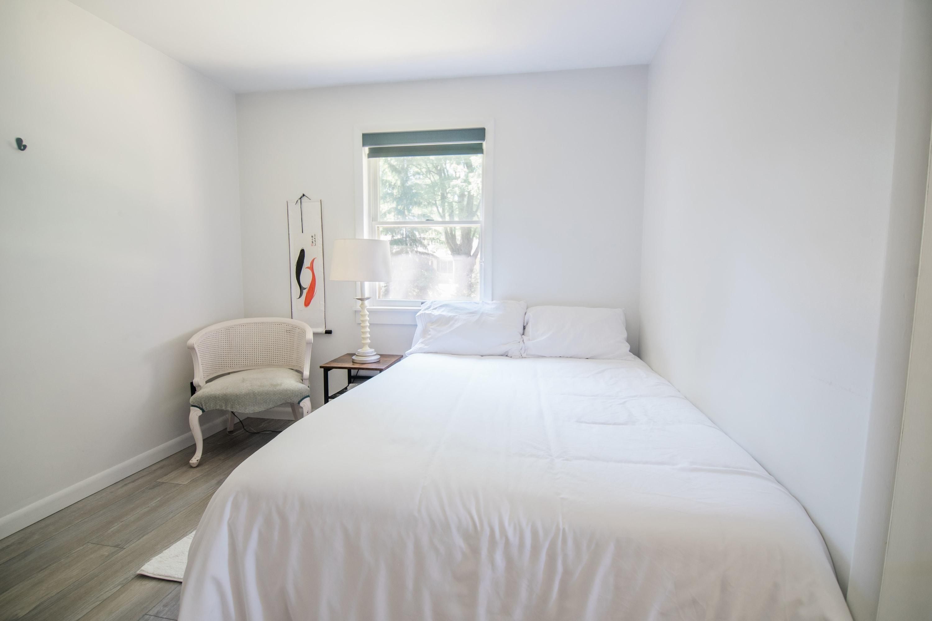 Un dormitorio con cama, una silla, una mesita de noche, una lámpara y una ventana.