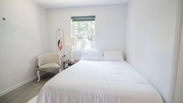 Un dormitorio con cama, una silla, una mesita de noche, una lámpara y una ventana.