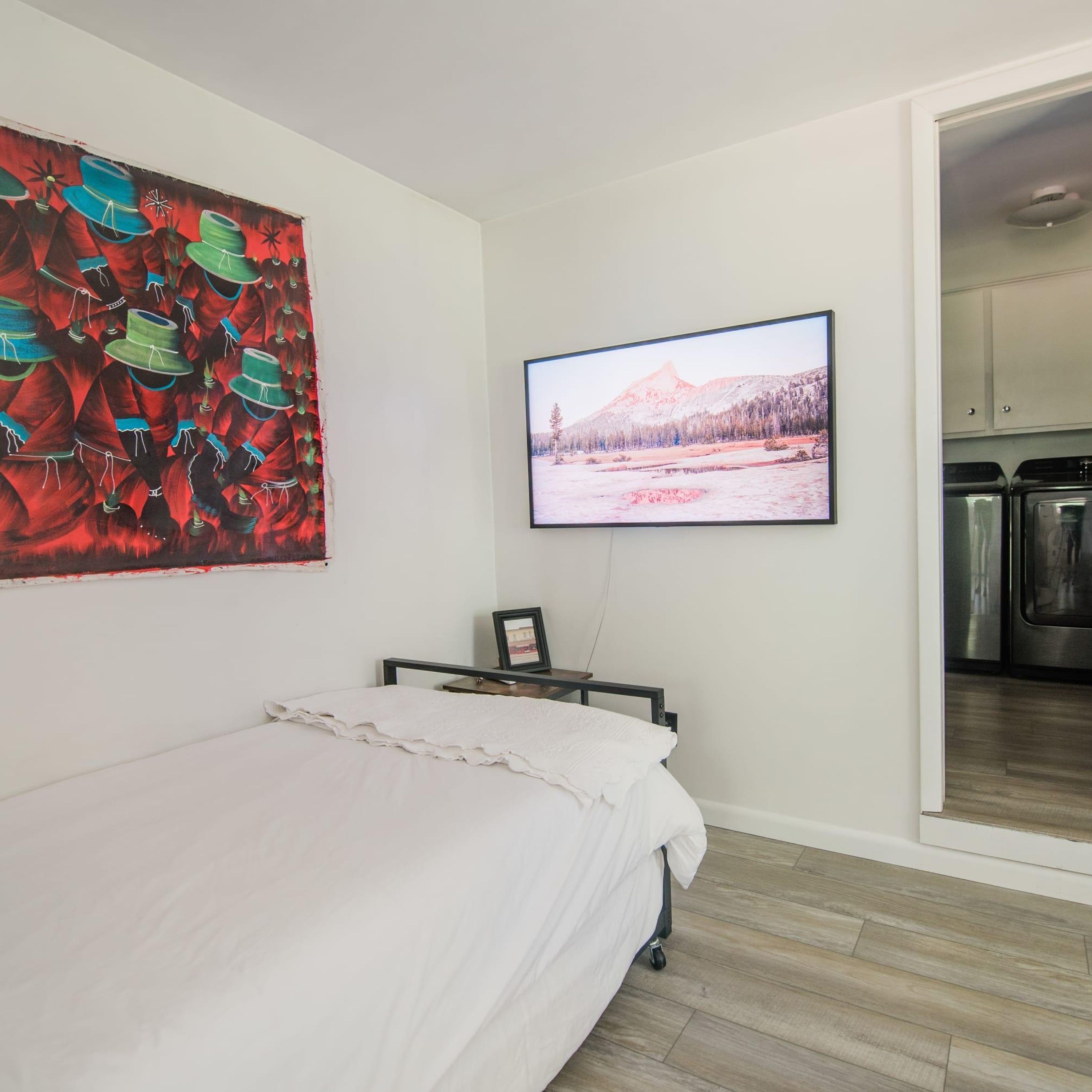 A bedroom with a bed, a painting, and a TV on the wall.
