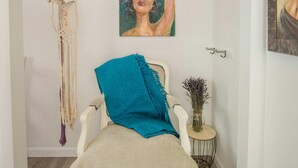 Ein Zimmer mit einem weißen Sessel, einem Wandbehang aus Makramee, einem gerahmten Gemälde und einem kleinen Beistelltisch mit einer Vase.