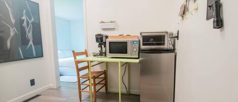 A compact kitchen with a microwave, toaster, and refrigerator. A chair is placed at a small table, and a large abstract painting is on the wall.