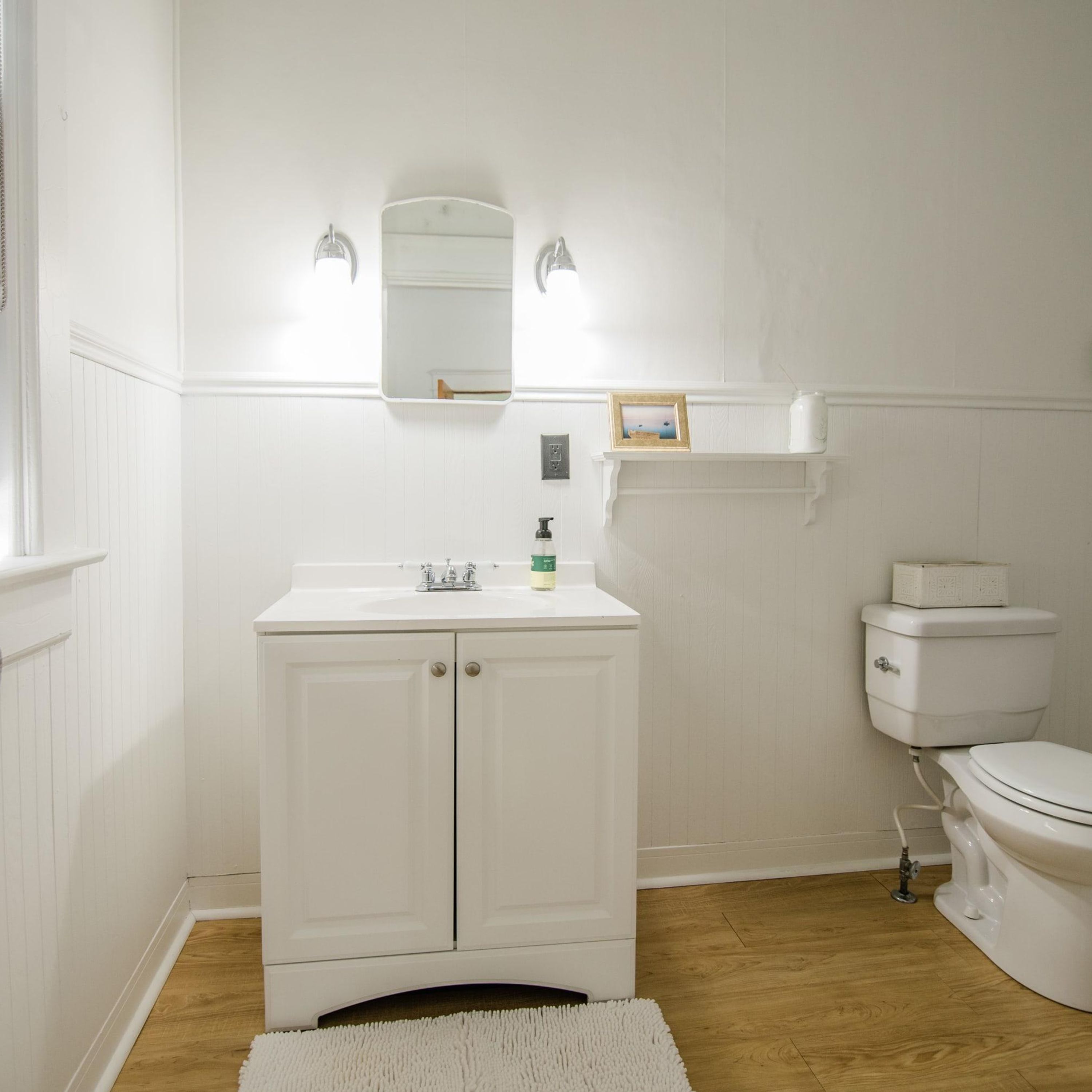 A bathroom with a white vanity, a mirror, a toilet, and a shelf above the toilet.